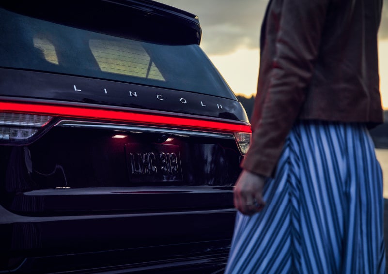 A person is shown near the rear of a 2024 Lincoln Aviator® SUV as the Lincoln Embrace illuminates the rear lights | Sheehy Lincoln of Gaithersburg in Gaithersburg MD